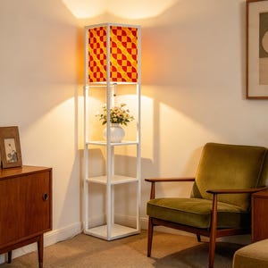 May include: A white floor lamp with a square frame and three open shelves. The top section features a fabric lampshade with a repeating orange and red geometric pattern. A small white vase with flowers sits on the middle shelf of the lamp.