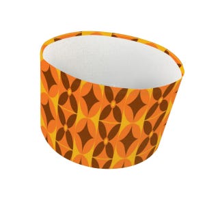 May include: A cylindrical lamp shade featuring a repeating geometric pattern of dark brown four-pointed stars centered within orange oval shapes, set against a yellow background. The interior of the lamp shade is lined with a plain white material.