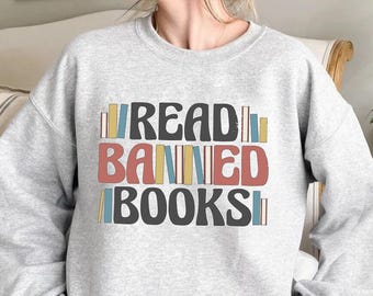 Pode incluir: Camisola cinza claro com o texto "READ BANNED BOOKS" em uma fonte retrô colorida. A palavra "READ" é preta, "BANNED" é vermelha e "BOOKS" é preta. A camisola tem gola redonda e mangas compridas.