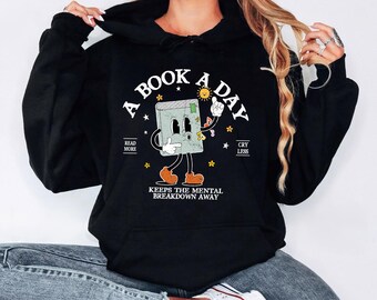 May include: Black hooded sweatshirt featuring the text "A BOOK A DAY" and a cartoon book character. The book character is light green with brown shoes and is holding a sun. The text "READ MORE" and "CRY LESS" are also on the sweatshirt.