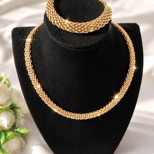 Könnte beinhalten: Ein goldfarbenes Schmuckset bestehend aus einer mehrsträngigen geflochtenen Kettenhalskette und einem passenden geflochtenen Kettenarmband. Beide Teile haben eine polierte metallische Oberfläche mit einem strukturierten, ineinandergreifenden Glieddesign.