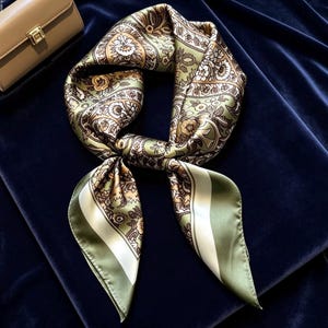 May include: A square silk scarf featuring a detailed floral and paisley pattern in shades of olive green, brown, and cream. The scarf is tied in a knot, displaying a wide solid olive green border along the edges and a smooth, lustrous finish.