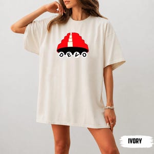 May include: An ivory short-sleeve t-shirt featuring a graphic of a red energy dome hat with a white vertical stripe. Below the hat, the word DEVO is printed in black capital letters, with each letter enclosed in a white circle.