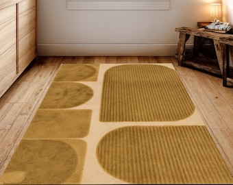 May include: A rectangular area rug with a geometric design in shades of tan and mustard yellow. The rug features rounded and rectangular shapes, with a textured, striped pattern on some sections. The rug is placed on a wooden floor.