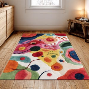May include: A vibrant, abstract patterned rug. The rug has a white base with large, overlapping shapes in red, pink, yellow, orange, green, and blue. It is rectangular and placed on a wooden floor, adding a splash of colour to the room.