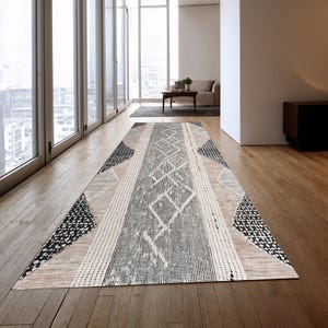 Op de afbeelding: Een rechthoekig vloerkleed met een geometrisch ontwerp. Het kleed heeft een centraal grijs gedeelte met ruitpatronen, begrensd door beige, zwarte en witte patronen. Het ligt op een houten vloer in een lichte kamer.