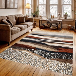 May include: A rectangular area rug with a textured, abstract design. The rug features horizontal bands of various colors, including brown, beige, gray, and black. The rug is placed on a hardwood floor in a living room setting.