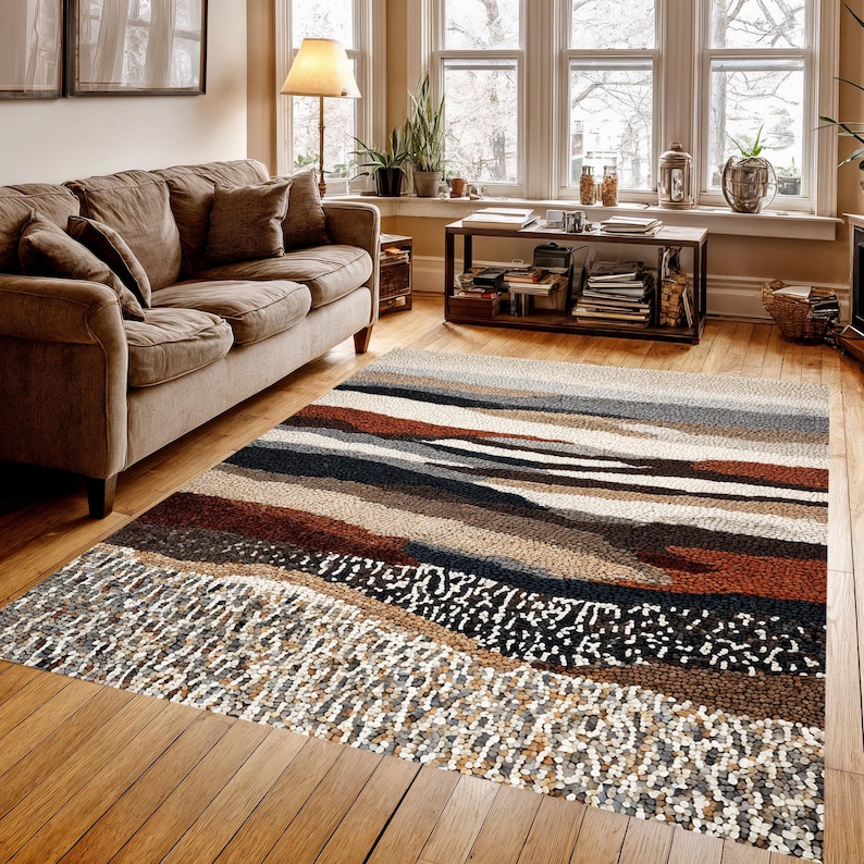 May include: A rectangular area rug with a textured, abstract design. The rug features horizontal bands of various colors, including brown, beige, gray, and black. The rug is placed on a hardwood floor in a living room setting.