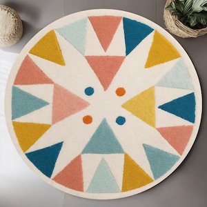 May include: A round, off-white rug with a geometric design. The pattern features triangles in shades of pink, yellow, teal, and blue, arranged around a central point. Small orange and blue dots accent the design.