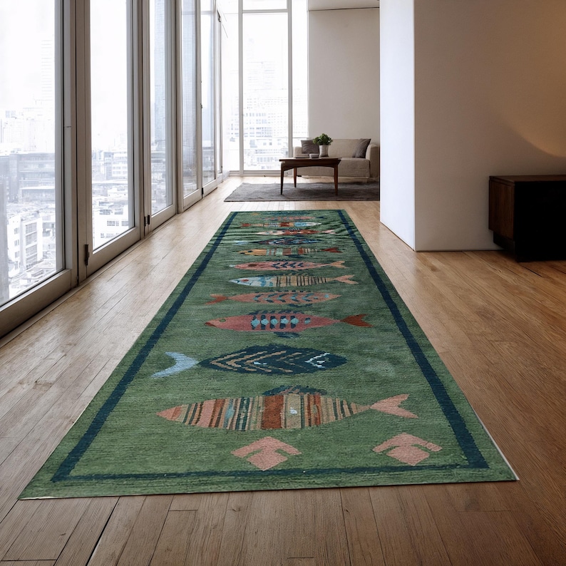 May include: A long, rectangular green rug with a dark blue border and a repeating pattern of colourful fish. The fish designs feature various colours and patterns, adding a whimsical touch to the floor covering. The rug is placed on a wooden floor.