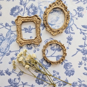May include: Three antique-style gold picture frames, square, oval, and round, are arranged on a patterned fabric. A small bunch of dried white flowers lies nearby. The fabric has a white base with a blue floral design.