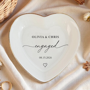 Puede incluir: Un plato de cerámica blanco en forma de corazón con borde dorado. El plato tiene la inscripción "OLIVIA & CHRIS engaged 08.15.2026" en escritura negra. Un pequeño símbolo de corazón está debajo de la fecha. El plato está rodeado de flores y un anillo dorado.