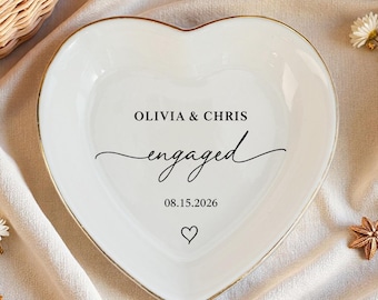 Könnte beinhalten: Eine herzförmige weiße Keramik-Ringschale mit goldenem Rand. Auf der Schale steht in schwarzer Schrift "OLIVIA & CHRIS engaged 08.15.2026". Unter dem Datum befindet sich ein kleines Herzsymbol. Die Schale ist von Blumen und einem goldenen Ring umgeben.