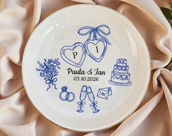 Puede incluir: Un plato de cerámica blanca con borde dorado que presenta ilustraciones de línea azul de dos relicarios en forma de corazón con las letras P e I, un pastel, un ramo, anillos, copas de champán y un sobre. El texto dice Paula & Ian 03.10.2026.
