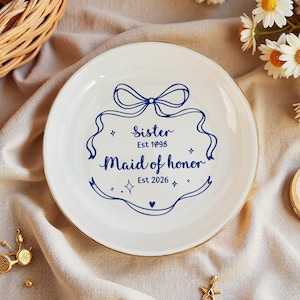 Puede incluir: Un plato de cerámica blanco con borde dorado, con texto azul que dice "Sister Est 1998 Maid of Honor Est 2026". El plato está decorado con un diseño de lazo y cinta. También hay un anillo y joyas doradas.