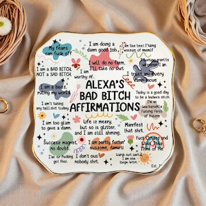 Könnte beinhalten: Eine weiße Keramik-Dekorplatte mit goldenem Rand, auf der in schwarzer Schrift "ALEXA'S BAD BITCH AFFIRMATIONS" steht, zusammen mit verschiedenen handgeschriebenen Affirmationen. Die Platte ist mit dekorativen Elementen verziert.