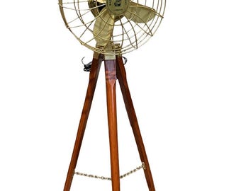 May include: A floor-standing electric fan featuring a brass-toned metal cage and blades mounted on a dark brown wooden tripod base. The center of the fan includes a circular emblem with the text Made for Royal Navy London.
