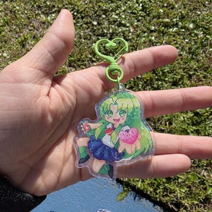 以下が含まれることがあります： 緑色の髪と目をしたアニメスタイルの漫画の女の子が学校の制服を着た、透明なアクリルキーホルダー。 キーホルダーには緑色のハート型の留め金とテキスト付きのピンクのハートが付いています。 背景はぼやけています。