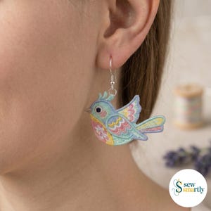 Puede incluir: Un pendiente en forma de pájaro de colores pastel, con un gancho plateado. El pájaro presenta un cuerpo azul, alas amarillas y detalles rosas y amarillos. El pendiente se lleva en la oreja de una persona.
