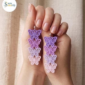 Puede incluir: Un par de pendientes colgantes con una cadena vertical de cuatro mariposas bordadas en un degradado de color, que va desde el morado oscuro en la parte superior hasta el rosa claro en la parte inferior, sujetos por ganchos de metal plateado.