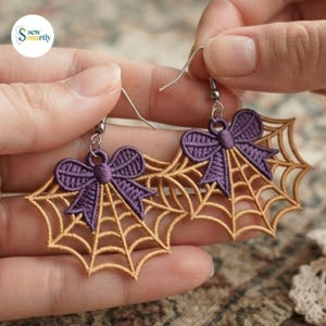 May include: A pair of dangle earrings featuring embroidered gold spider web motifs topped with purple bows. The earrings are attached to silver-toned metal hooks. The design is intricate with detailed stitching forming the web pattern and the bow shape.