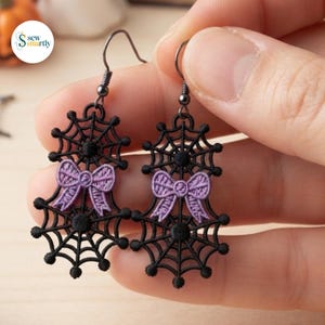 May include: A pair of black embroidered spider web earrings featuring a small lavender bow at the center of each web. The earrings are attached to dark metal fishhook wires and showcase intricate thread work in a circular web pattern.