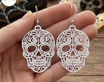 Puede incluir: Un par de pendientes blancos con forma de calavera que presentan intrincados patrones de filigrana recortados. Los pendientes están fabricados con un material ligero de textura similar al encaje y unidos a ganchos de metal plateado.