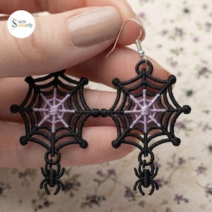May include: A pair of dangling earrings featuring black embroidered spider web motifs with light purple centers. Each web is connected to a small black spider charm at the bottom. The earrings are attached to silver-toned metal fishhook wires.