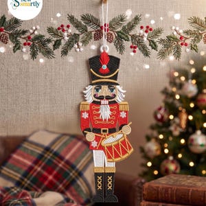 Puede incluir: Adorno navideño bordado de un cascanueces rojo, negro y dorado. El adorno está suspendido por un hilo blanco. El cascanueces lleva un sombrero negro con una pluma roja y sostiene un tambor. El fondo presenta un árbol de Navidad y una guirnalda festiva.