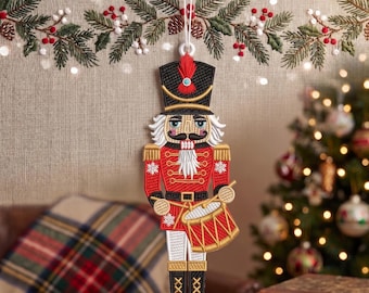以下が含まれることがあります： 赤、黒、金色の刺繍が施されたくるみ割り人形のクリスマスオーナメント。白い紐で吊るされています。くるみ割り人形は赤い羽根飾りのついた黒い帽子をかぶり、太鼓を持っています。背景にはクリスマスツリーとガーランドがあります。