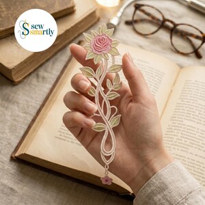 Può includere: Un segnalibro floreale con una rosa rosa e foglie verdi. Il segnalibro è tenuto sopra un libro aperto. Il segnalibro ha un design delicato, simile al pizzo. Il logo "Sew Smartly" si trova nell'angolo in alto a sinistra.