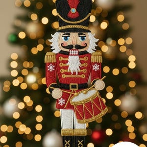 Könnte beinhalten: Gestickter Weihnachtsschmuck eines Nussknackersoldaten. Der Schmuck hat eine rote Jacke, einen schwarzen Hut und weiße Haare und einen Schnurrbart. Er hält eine rote Trommel mit goldenen Akzenten. Der Hintergrund ist ein verschwommener Weihnachtsbaum mit Bokeh-Lichtern.