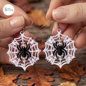May include: A pair of white thread-embroidered spider web earrings, each featuring a centered black thread spider. The earrings are suspended from silver-toned metal hooks and display intricate, circular web patterns with radiating spokes.
