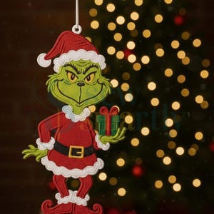 May include: Embroidered Christmas ornament featuring the Grinch in a Santa suit, holding a small gift. The Grinch is green with yellow eyes and a grumpy expression. The ornament is red and white with a black belt. A Christmas tree with bokeh lights is in the background.