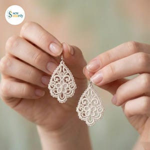 Pode incluir: Um par de brincos de renda em forma de lágrima, na cor branco-sujo, com um intrincado padrão de bordado floral. Cada brinco está suspenso por um gancho de metal prateado, exibindo um delicado design rendilhado com um motivo floral central.