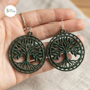 May include: A pair of circular dark green embroidered earrings featuring a detailed tree of life design. Each earring is suspended from a silver-toned metal hook and showcases intricate thread work forming branches, leaves, and roots within a circular frame.