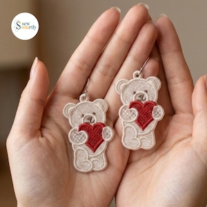 May include: A pair of embroidered dangle earrings featuring cream-colored teddy bears, each holding a bright red heart. The bears have stitched facial details and textured fur patterns, suspended from silver-toned metal earring hooks.