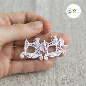 May include: A pair of white lace earrings shaped like vintage sewing machines. The earrings feature intricate openwork embroidery detailing the machine frame, needle bar, and base, suspended from silver-toned metal ear wires for pierced ears.