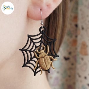 May include: A dangling earring featuring a black embroidered spider web design with a golden-brown beetle stitched in the center. The piece is suspended from a silver-toned metal hook and showcases intricate thread work throughout the entire shape.
