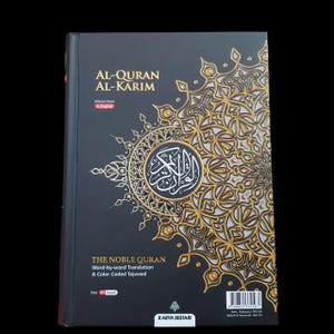 May include: A hardcover book titled "AL-QURAN AL-KARIM" in English. The cover is black with gold filigree and a central circular emblem. The text "THE NOBLE QURAN" and "Word-by-word Translation & Color Coded Tajweed" are also visible.