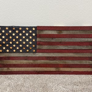 May include: Wooden American flag wall art. The flag features a black field with star cutouts and red and gray stripes. The flag is made of reclaimed wood and is a rustic, patriotic decoration.