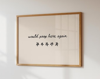May include: A framed print with a light beige background and a wooden frame. The black text reads "would poop here again" with five star symbols below. The print is displayed on a white wall.