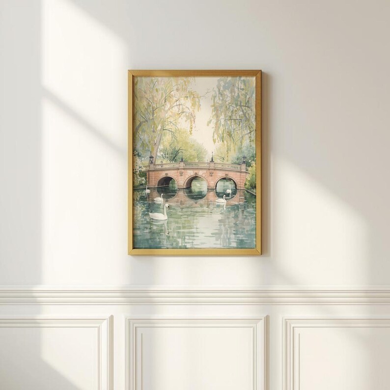 May include: A framed watercolor painting of a serene park scene. A stone bridge with three arches spans a calm body of water, with several white swans swimming. The artwork is in a gold frame and hangs on a white wall.