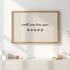 May include: A framed print with the text "would poop here again" in black script, along with five star symbols. The print has a light beige background and a natural wood frame. The artwork is displayed on a white wall.