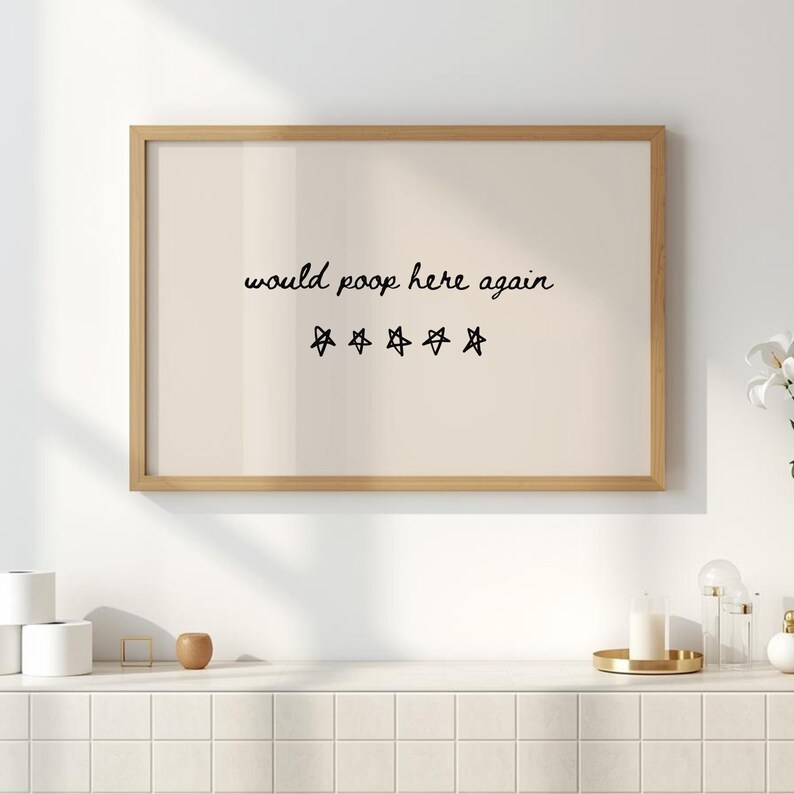 May include: A framed print with the text "would poop here again" in black script, along with five star symbols. The print has a light beige background and a natural wood frame. The artwork is displayed on a white wall.