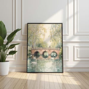 May include: A framed watercolor painting depicts a serene waterscape with a stone bridge and swans. The bridge has three arches and is surrounded by lush greenery and weeping trees. The artwork is displayed on a wooden floor.