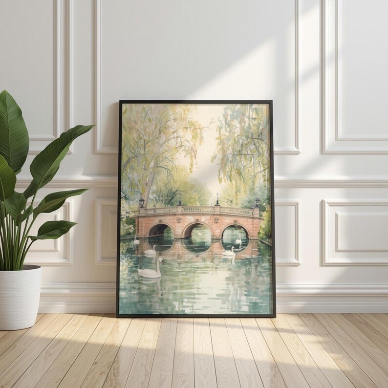 May include: A framed watercolor painting depicts a serene waterscape with a stone bridge and swans. The bridge has three arches and is surrounded by lush greenery and weeping trees. The artwork is displayed on a wooden floor.