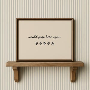May include: A framed print with the text "would poop here again" and five star symbols. The frame is brown, and the print is displayed on a wooden shelf. The background features a striped wallpaper.