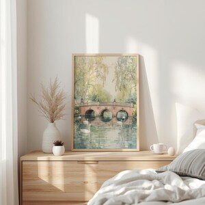 May include: A framed watercolor print of a bridge over a lake with swans. The bridge is made of red brick and has three arches. The water reflects the bridge and the surrounding trees. The print is in a light wood frame.