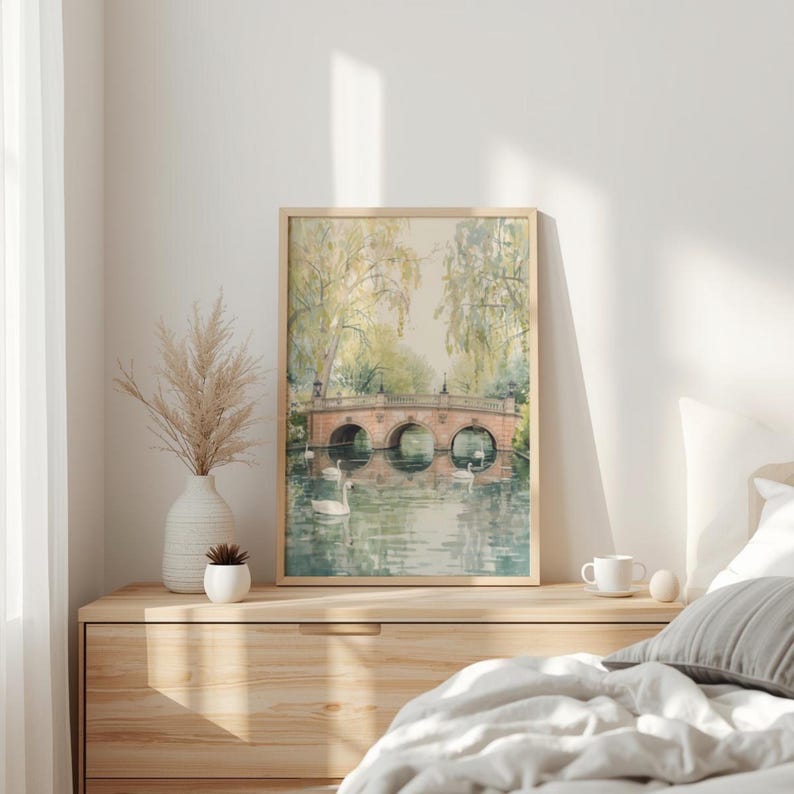 May include: A framed watercolor print of a bridge over a lake with swans. The bridge is made of red brick and has three arches. The water reflects the bridge and the surrounding trees. The print is in a light wood frame.